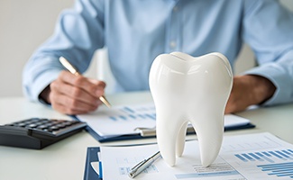 Man in blue shirt calculating with pen, paper, and calculator at desk with large model tooth